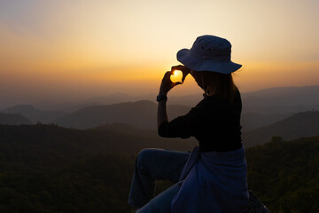 Wall Mural - Silhouette of woman making a heart shape with her hands in front of a sunset. Symbol of love. Manifestation of love. Expression of feelings. Happiness, youth.