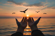 Hands outstretched upwards in a gesture of worship against a serene sunset over water, with birds soaring above. Symbolizing prayer and seeking divine blessings