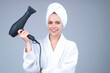 Woman in bathrobe and towel on head with hairdryer in studio. Portrait of female model with blow dryer. Hair care and beauty. Morning routine.