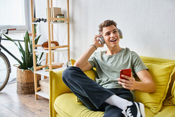 Wall Mural - charming man with headphones in yellow couch at home scrolling to social media and looking to side