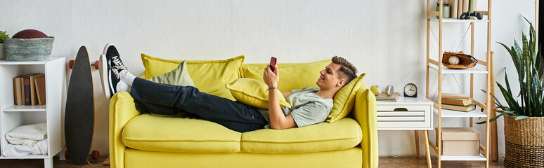 Wall Mural - horizontal shot of young man lying on yellow couch in living room and scrolling to social media