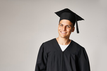 Wall Mural - portrait of cheerful young student in graduate gown and cap with grey eyes against light background