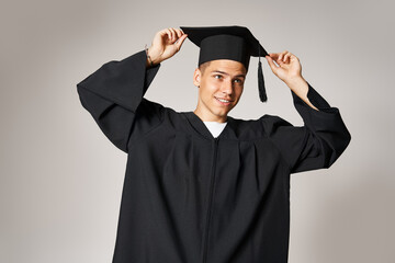 Wall Mural - attractive student in gown with grey eyes holding graduate cap on head in light background