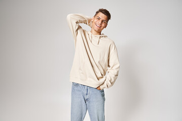 Wall Mural - charming man in white hoodie smiling and putting hand behind head against light background