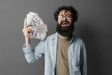 bearded man in casual attire showing excitement while holding a fan of dollar bills against a plain 