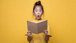 A shocked Asian girl is reading a brown book on a plain yellow background with copy-space for text.