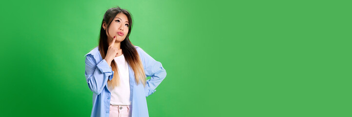 Banner. Thoughtful woman holding hand on chin against vibrant green studio background with negative space to insert text. Concept of human emotions, self-expression, fashion, style.