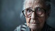 Thoughtful elderly woman looking at the camera with a serious expression on her face. She is wearing glasses and has gray hair.