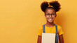 A happy and smiling black girl wearing glasses is holding a white book on a plain yellow background with copy-space for text.