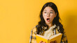 A shocked Asian teenage girl wearing glasses is reading a yellow book on a plain green background with copy-space for text.