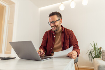A happy adult businessman with glasses looking at his finances on a laptop while working from home