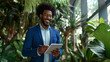 Homem afro vestindo um terno azul segurando um tablet em um escritório cheio de plantas - Conceito de sustentabilidade
