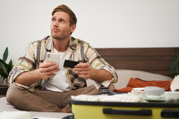 Portrait of young man booking tickets, going on holiday, holding credit card and smartphone, sitting with mobile phone near suitcase filled with clothes, ready for vacation trip