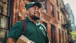 A man in a green shirt and cap smiling and carrying a box walking down a city street with brick buildings.