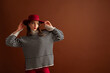Fashionable confident woman wearing oversized striped knitted turtleneck sweater with wide sleeves, red hat, posing on brown background. Studio fashion portrait. Copy, empty, blank space for text