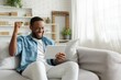 An African-American men sitting on a sofa and holding a tablet rejoices at success.