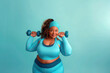 A joyful plus-size sportswoman wearing a blue sports bra top, holding a pair of dumbbells.
