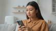 © Yuliia - Korean chinese multiracial woman use smartphone application surfing internet social media pensive Asian housewife girl in kitchen typing mobile phone message buy goods online order delivery at home