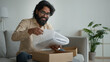 © Yuliia - Happy male customer client consumer Arabian indian man guy in glasses open cardboard box at home unpack online shopping order purchase wrapped package get delivery present gift unpacking carton parcel