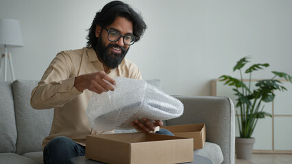 Happy male customer client consumer Arabian indian man guy in glasses open cardboard box at home unpack online shopping order purchase wrapped package get delivery present gift unpacking carton parcel