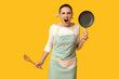 Portrait of angry young housewife in apron with frying pan and wooden spatula on yellow background