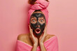 surprised attractive girl applies a black cleansing mask on her face, on her head a pink towel