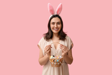 Wall Mural - Beautiful woman in bunny ears holding Easter basket with decorative cosmetics on pink background