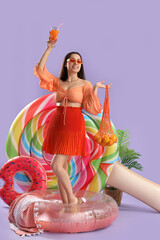 Sticker - Smiling young woman in sunglasses holding cocktail with lemons, inflatable rings and mattress on lilac background