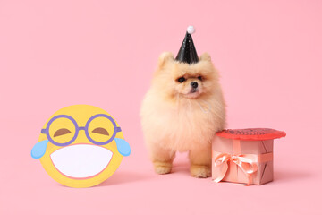 Wall Mural - Cute Pomeranian Spitz dog in party hat with paper smile and gift box on pink background. April Fools day celebration