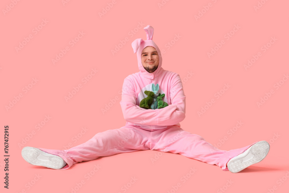 Young man in bunny costume with toy rabbits sitting on pink background. Easter celebration