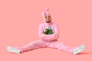 Wall Mural - Young man in bunny costume with toy rabbits sitting on pink background. Easter celebration