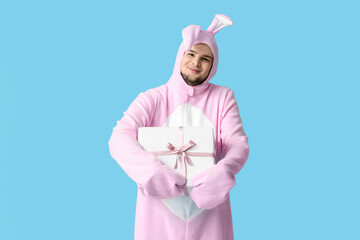 Wall Mural - Young man in bunny costume with gift box on blue background. Easter celebration