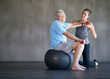 © YATrainer/peopleimages.com - Physiotherapist, dumbbells and smile of senior man on ball for fitness or rehabilitation at gym on mockup. Elderly person, weightlifting and personal trainer help for body health or physical therapy