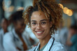© Nataliia_Trushchenko - A woman with curly hair and glasses is smiling. She is wearing a white lab coat and is posing for a picture. smiling afro doctor with colleagues in background, looking at camera