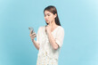 Beautiful young asian woman in white dress with flower pattern using smartphone with unsure feeling isolated on blue background