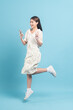 Beautiful young asian woman in white dress with flower pattern using smartphone and jump  isolated on blue background