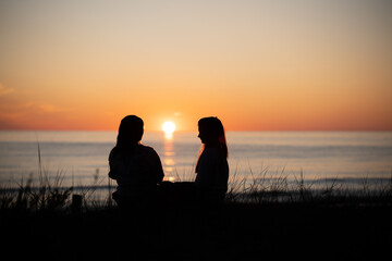 Silhouette of two female friends enjoying a conversation while sitting and watching the sunset on a sea beach. Back view