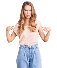 Sticker - Young beautiful caucasian woman with blond hair wearing casual clothes pointing down looking sad and upset, indicating direction with fingers, unhappy and depressed.