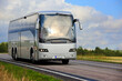 Silver coach bus traveling along highway through rural scenery in late summer, blue sky and clouds in the background.