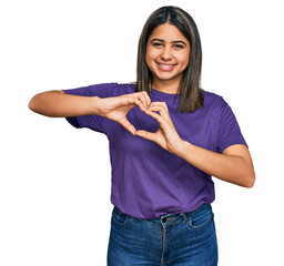 Poster - Young hispanic girl wearing casual purple t shirt smiling in love doing heart symbol shape with hands. romantic concept.