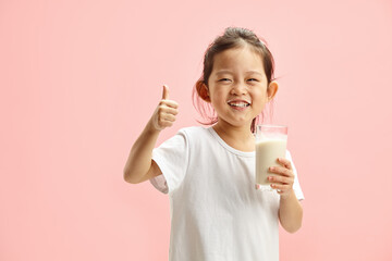 Wall Mural - Happy Smiling Little Brunette Child Girl Drinking Milk From Glass With a Trace Frm Milk on Lips, Expresses Pleasure Positively Showing Thumb up Standing On Pink Isolated