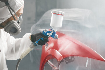 Wall Mural - Worker in protective mask and jumpsuit overall spraying base coat of paint on car body elements in painted chamber using professional airbrush.