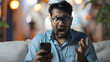 Indian guy staring at smartphone screen with angry face expression, get awful news in notice from bank, bad message, scam, fraud, screams due to short battery life, lagging phone, mobile apps crash