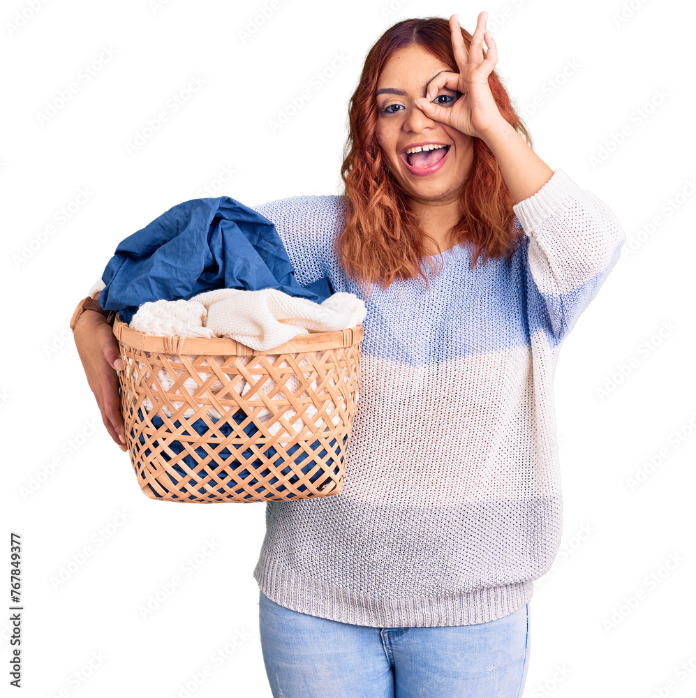 Young latin woman holding laundry basket smiling happy doing ok sign with hand on eye looking through fingers