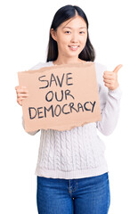 Wall Mural - Young beautiful chinese woman holding save our democracy cardboard banner smiling happy and positive, thumb up doing excellent and approval sign