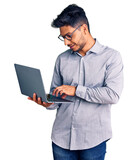 Fototapeta Panele - Handsome latin american young man working using computer laptop looking positive and happy standing and smiling with a confident smile showing teeth