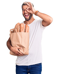 Canvas Print - Young handsome blond man holding paper bag with bread smiling happy doing ok sign with hand on eye looking through fingers