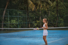 A Beautiful Girl With Long Blond Hair Gathered In A Ponytail Plays Tennis. Tennis Court. A Girl Holds A Tennis Racket And A Tennis Ball. A Game. Sport. Tennis Court On The Island, Among Palm Trees.