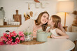 Child congratulates mother with flowers on Mother's Day. Family communication of mom and kid. Happy mother with daughter drinking tea or cocoa sitting in kitchen and having fun. Spend time together.