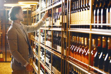 Sommelier stands in cellar and taking out one wine of many submitted from wooden shelf. Male bartender in wine storage choose necessary alcoholic bottle for buyer based on his requests.